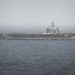 A large U.S. Navy aircraft carrier bearing hull number '73' is underway at sea under overcast, hazy conditions. The flight deck is populated with multiple aircraft, and at least one aircraft appears to be launching or departing from the bow. The vessel is moving through calm, dark ocean waters with a visible wake at the stern.