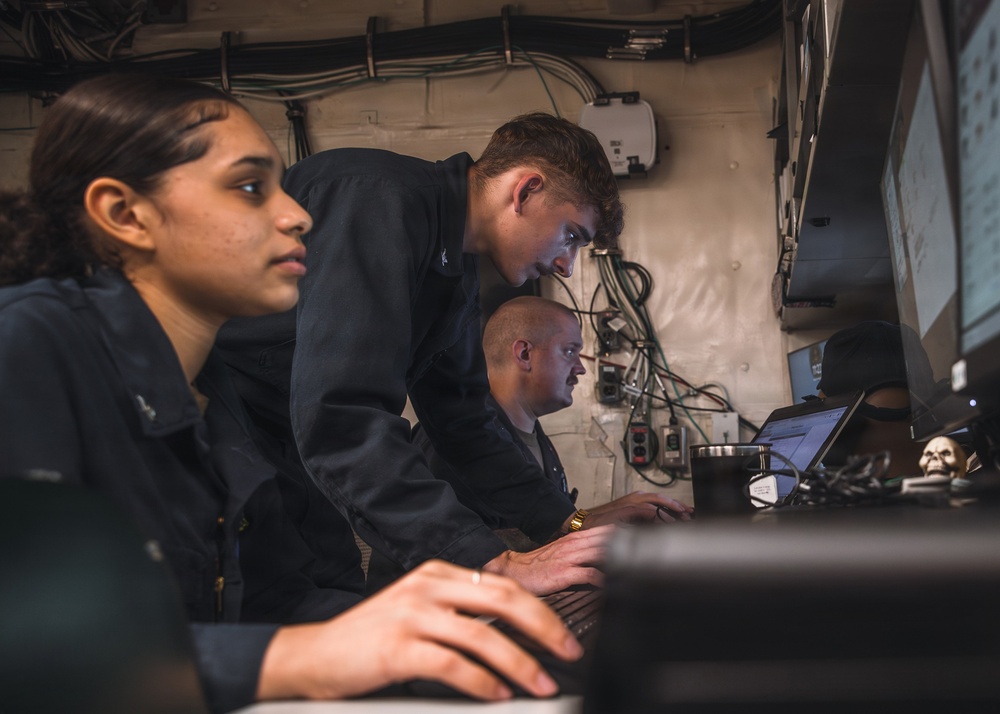 Three individuals wearing matching dark navy/military-style uniforms are working together at a workstation with laptops and monitors in what appears to be a confined technical or operations space. The setting features exposed wiring, cable management systems, and mounted equipment on the walls, consistent with a military vessel or operations center. Two individuals are seated while one leans over to observe or assist with the work on screen.