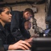 Three individuals wearing matching dark navy/military-style uniforms are working together at a workstation with laptops and monitors in what appears to be a confined technical or operations space. The setting features exposed wiring, cable management systems, and mounted equipment on the walls, consistent with a military vessel or operations center. Two individuals are seated while one leans over to observe or assist with the work on screen.