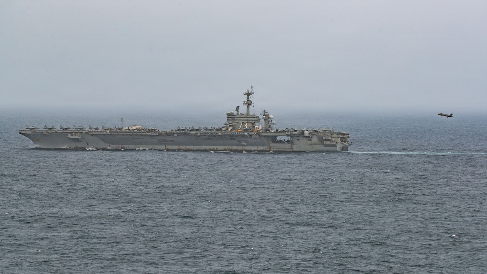 A large US Navy aircraft carrier bearing hull number '73' is underway at sea under overcast, hazy conditions. Multiple aircraft are visible parked on the flight deck, and a single jet aircraft is seen airborne to the right of the vessel, likely having just launched or conducting a flyby. The ship appears to be the USS George Washington (CVN-73), a Nimitz-class nuclear-powered supercarrier.
