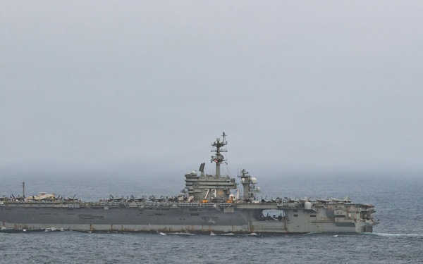 A large US Navy aircraft carrier bearing hull number '73' is underway at sea under overcast, hazy conditions. Multiple aircraft are visible parked on the flight deck, and a single jet aircraft is seen airborne to the right of the vessel, likely having just launched or conducting a flyby. The ship appears to be the USS George Washington (CVN-73), a Nimitz-class nuclear-powered supercarrier.