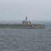 A large US Navy aircraft carrier bearing hull number '73' is underway at sea under overcast, hazy conditions. Multiple aircraft are visible parked on the flight deck, and a single jet aircraft is seen airborne to the right of the vessel, likely having just launched or conducting a flyby. The ship appears to be the USS George Washington (CVN-73), a Nimitz-class nuclear-powered supercarrier.