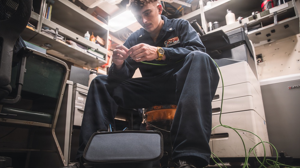 A young male technician wearing dark navy coveralls is seated in a confined workspace, working with green wiring or cabling. He appears to be in a military or industrial environment, likely aboard a naval vessel, based on his uniform and the cluttered technical workspace surrounding him. A gold wristwatch is visible on his wrist as he manipulates the green wire with both hands.