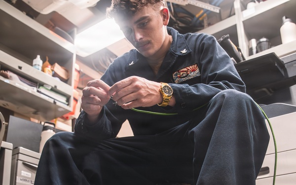 A young male technician wearing dark navy coveralls is seated in a confined workspace, working with green wiring or cabling. He appears to be in a military or industrial environment, likely aboard a naval vessel, based on his uniform and the cluttered technical workspace surrounding him. A gold wristwatch is visible on his wrist as he manipulates the green wire with both hands.