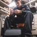 A young male technician wearing dark navy coveralls is seated in a confined workspace, working with green wiring or cabling. He appears to be in a military or industrial environment, likely aboard a naval vessel, based on his uniform and the cluttered technical workspace surrounding him. A gold wristwatch is visible on his wrist as he manipulates the green wire with both hands.