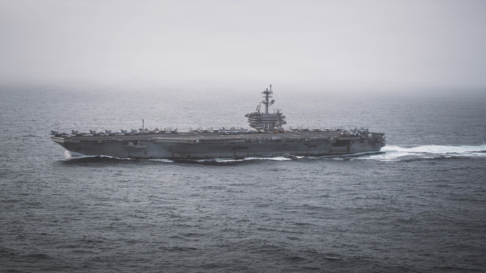 A large US Navy aircraft carrier is underway at sea under overcast, hazy conditions. The hull number '73' is clearly visible on the island superstructure, identifying it as the USS George Washington (CVN-73), a Nimitz-class nuclear-powered carrier. Multiple fixed-wing aircraft are visible parked on the flight deck.