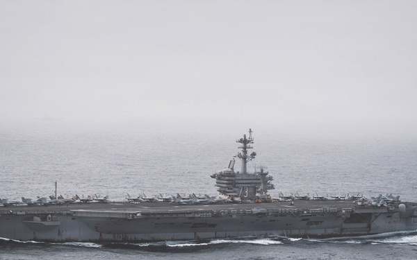 A large US Navy aircraft carrier is underway at sea under overcast, hazy conditions. The hull number '73' is clearly visible on the island superstructure, identifying it as the USS George Washington (CVN-73), a Nimitz-class nuclear-powered carrier. Multiple fixed-wing aircraft are visible parked on the flight deck.