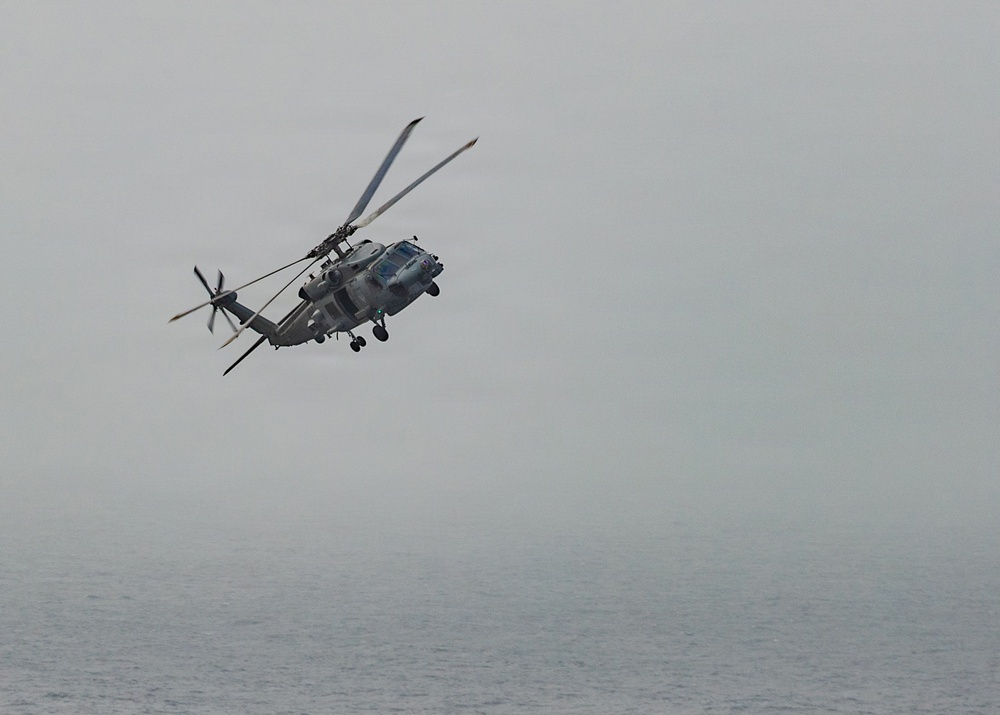 A military helicopter is captured in flight over a body of water under overcast, hazy conditions. The aircraft appears to be a naval helicopter, likely an SH-60 Seahawk or similar variant, flying at low altitude above the sea surface. The muted gray tones of both the sky and water create a monochromatic, atmospheric scene.