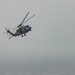 A military helicopter is captured in flight over a body of water under overcast, hazy conditions. The aircraft appears to be a naval helicopter, likely an SH-60 Seahawk or similar variant, flying at low altitude above the sea surface. The muted gray tones of both the sky and water create a monochromatic, atmospheric scene.