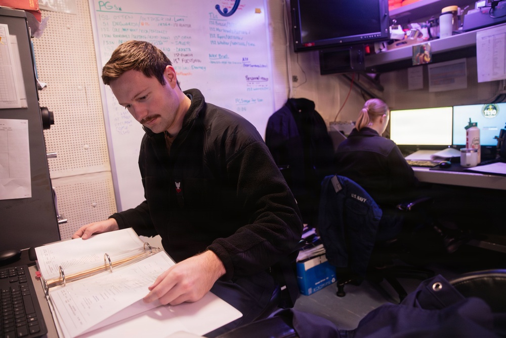 Two individuals are working in what appears to be a U.S. Navy operations or administrative workspace. The foreground person, a male with a mustache wearing a black fleece, is reviewing documents in a three-ring binder. A second person in the background, wearing a black jacket with 'U.S. NAVY' visible on a nearby bag, is working at a dual-monitor computer workstation.