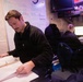 Two individuals are working in what appears to be a U.S. Navy operations or administrative workspace. The foreground person, a male with a mustache wearing a black fleece, is reviewing documents in a three-ring binder. A second person in the background, wearing a black jacket with 'U.S. NAVY' visible on a nearby bag, is working at a dual-monitor computer workstation.