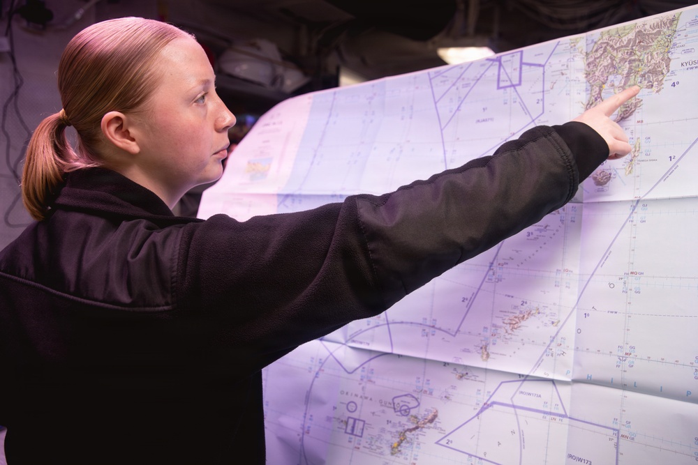 A young woman with red hair in a ponytail, wearing a black fleece jacket, is pointing at a large nautical or aeronautical chart. The map appears to show the Kyushu region of Japan and surrounding waters, including Okinawa. She appears to be in a dimly lit operational or military environment.