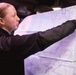 A young woman with red hair in a ponytail, wearing a black fleece jacket, is pointing at a large nautical or aeronautical chart. The map appears to show the Kyushu region of Japan and surrounding waters, including Okinawa. She appears to be in a dimly lit operational or military environment.