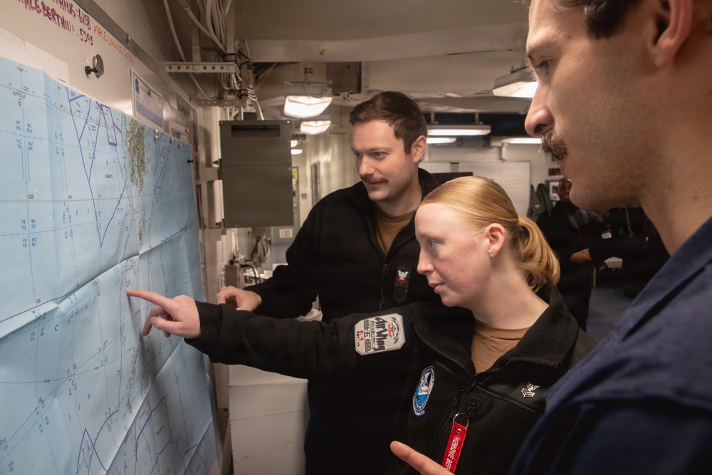 Three military personnel in black uniforms are gathered around a nautical or navigational chart mounted on a wall inside what appears to be a naval vessel. A woman in the center is pointing at a specific location on the map while two male colleagues observe. The setting appears to be a ship's navigation or operations room.