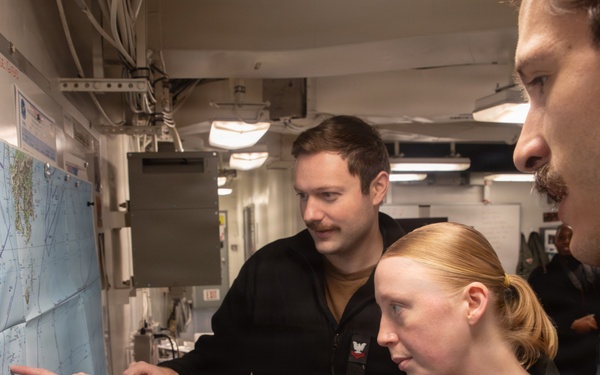 Three military personnel in black uniforms are gathered around a nautical or navigational chart mounted on a wall inside what appears to be a naval vessel. A woman in the center is pointing at a specific location on the map while two male colleagues observe. The setting appears to be a ship's navigation or operations room.