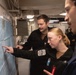 Three military personnel in black uniforms are gathered around a nautical or navigational chart mounted on a wall inside what appears to be a naval vessel. A woman in the center is pointing at a specific location on the map while two male colleagues observe. The setting appears to be a ship's navigation or operations room.