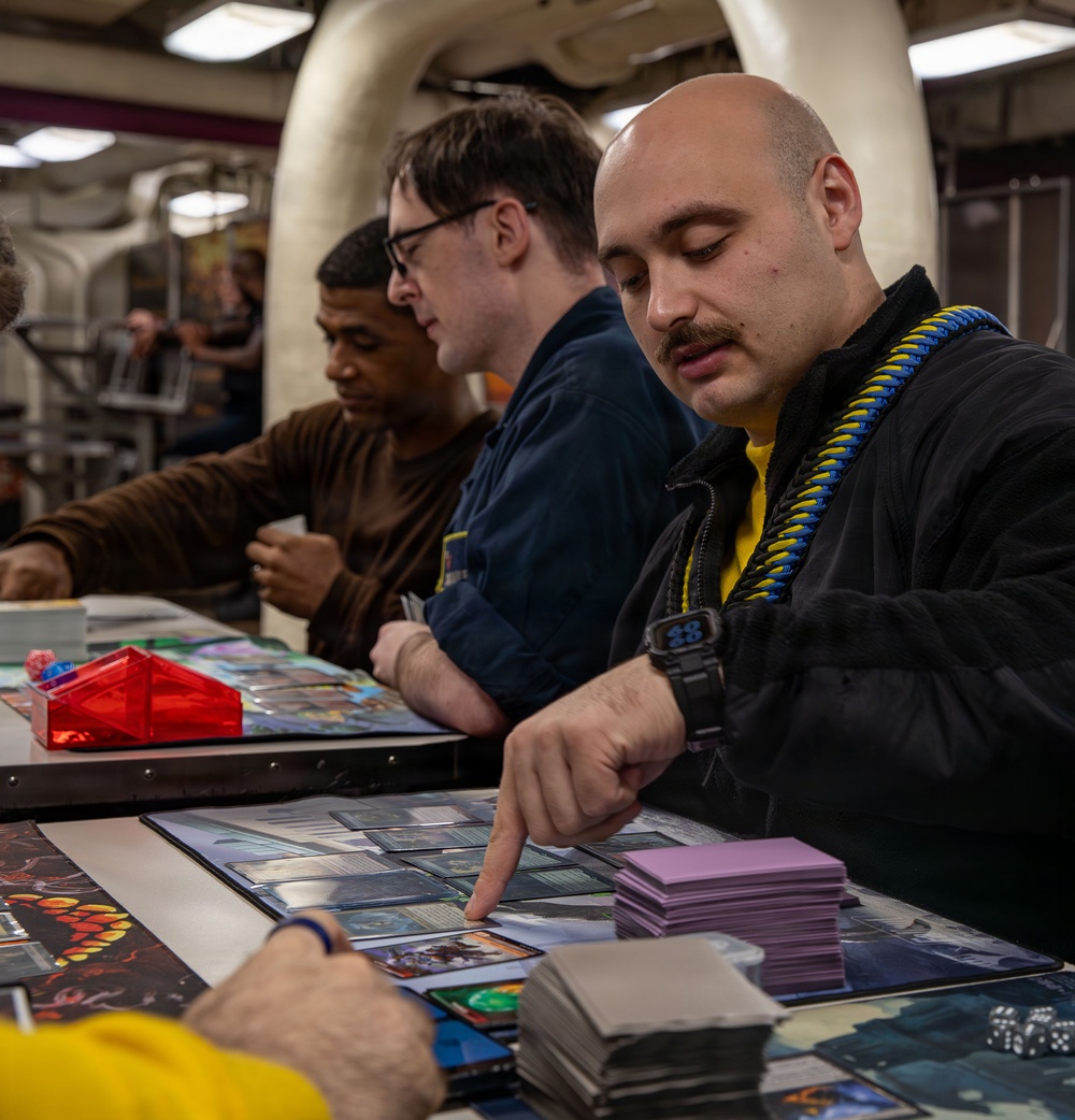 A group of people are engaged in a trading card game, likely Magic: The Gathering, at a table covered with game mats, cards, and accessories. The foreground player, a bald man with a mustache wearing a black jacket with a blue-and-yellow lanyard, is pointing at cards on the play mat. The setting appears to be an organized gaming event or tournament held in an industrial or warehouse-style venue.