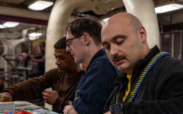 A group of people are engaged in a trading card game, likely Magic: The Gathering, at a table covered with game mats, cards, and accessories. The foreground player, a bald man with a mustache wearing a black jacket with a blue-and-yellow lanyard, is pointing at cards on the play mat. The setting appears to be an organized gaming event or tournament held in an industrial or warehouse-style venue.