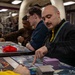 A group of people are engaged in a trading card game, likely Magic: The Gathering, at a table covered with game mats, cards, and accessories. The foreground player, a bald man with a mustache wearing a black jacket with a blue-and-yellow lanyard, is pointing at cards on the play mat. The setting appears to be an organized gaming event or tournament held in an industrial or warehouse-style venue.
