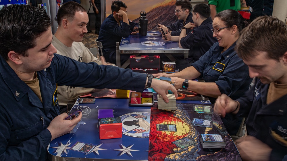 A group of individuals in matching dark blue uniforms are gathered around tables playing what appears to be a trading card game, likely Magic: The Gathering. The tables feature decorative playmats with patriotic and fantasy artwork. The setting appears to be a recreational or common area aboard a military vessel or installation.