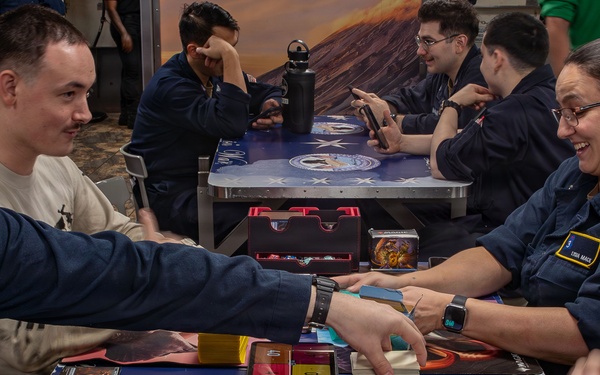A group of individuals in matching dark blue uniforms are gathered around tables playing what appears to be a trading card game, likely Magic: The Gathering. The tables feature decorative playmats with patriotic and fantasy artwork. The setting appears to be a recreational or common area aboard a military vessel or installation.