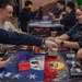 A group of individuals in matching dark blue uniforms are gathered around tables playing what appears to be a trading card game, likely Magic: The Gathering. The tables feature decorative playmats with patriotic and fantasy artwork. The setting appears to be a recreational or common area aboard a military vessel or installation.