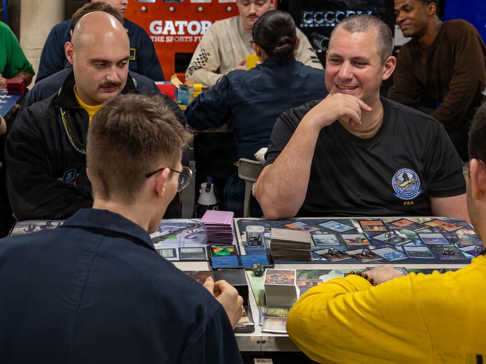 A group of people are engaged in a trading card game, likely Magic: The Gathering, at what appears to be a organized tournament or gaming event. The table is covered with playing cards, dice, and card sleeves. One player wearing a dark shirt with an 'FSA' logo is smiling confidently while facing an opponent across the table.