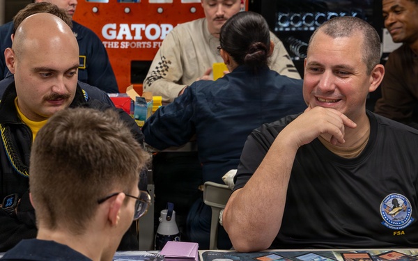 A group of people are engaged in a trading card game, likely Magic: The Gathering, at what appears to be a organized tournament or gaming event. The table is covered with playing cards, dice, and card sleeves. One player wearing a dark shirt with an 'FSA' logo is smiling confidently while facing an opponent across the table.