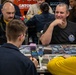 A group of people are engaged in a trading card game, likely Magic: The Gathering, at what appears to be a organized tournament or gaming event. The table is covered with playing cards, dice, and card sleeves. One player wearing a dark shirt with an 'FSA' logo is smiling confidently while facing an opponent across the table.