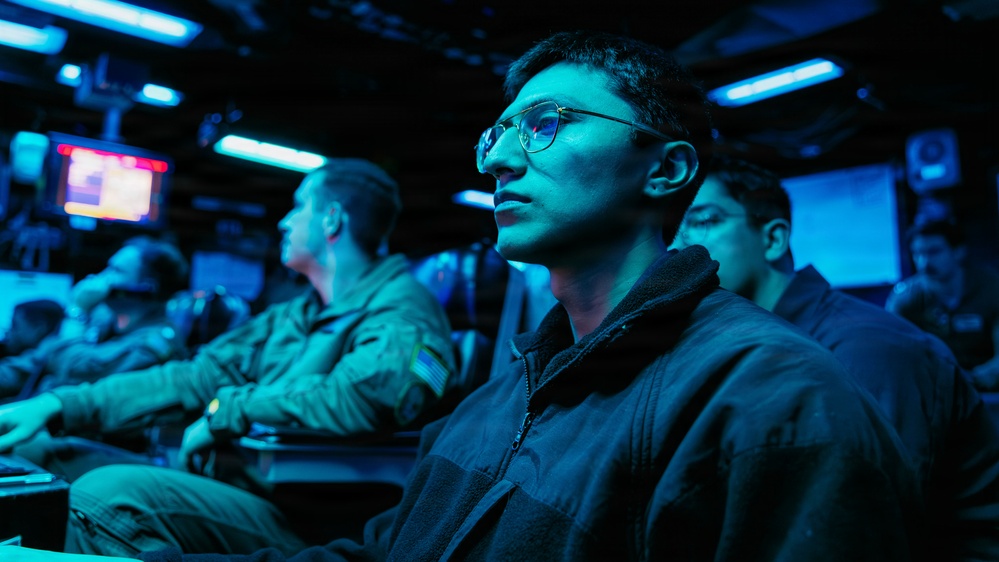 The image shows multiple individuals seated in a dimly lit room illuminated by blue ambient lighting, consistent with a military operations center, briefing room, or command and control environment. The foreground subject is a young person wearing glasses and a dark fleece jacket, appearing attentive and focused. Several other individuals in what appear to be military-style uniforms are visible in the background.