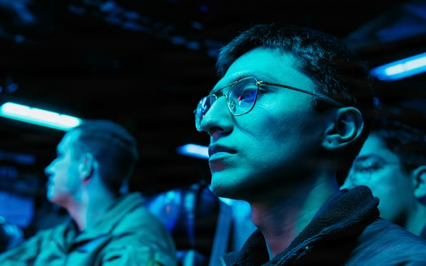 The image shows multiple individuals seated in a dimly lit room illuminated by blue ambient lighting, consistent with a military operations center, briefing room, or command and control environment. The foreground subject is a young person wearing glasses and a dark fleece jacket, appearing attentive and focused. Several other individuals in what appear to be military-style uniforms are visible in the background.