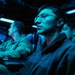 The image shows multiple individuals seated in a dimly lit room illuminated by blue ambient lighting, consistent with a military operations center, briefing room, or command and control environment. The foreground subject is a young person wearing glasses and a dark fleece jacket, appearing attentive and focused. Several other individuals in what appear to be military-style uniforms are visible in the background.