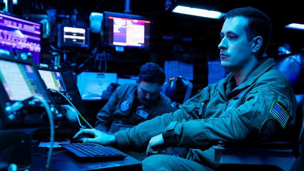 Two individuals in military flight suits are working at a multi-monitor operations center bathed in blue ambient lighting. The foreground operator is actively using a laptop while monitoring multiple screens displaying data and interface readouts. The environment appears to be a command, control, or intelligence operations facility.
