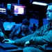 Two individuals in military flight suits are working at a multi-monitor operations center bathed in blue ambient lighting. The foreground operator is actively using a laptop while monitoring multiple screens displaying data and interface readouts. The environment appears to be a command, control, or intelligence operations facility.