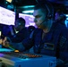 A U.S. Navy sailor named Brandon Palmer is seated at a combat or operations control console aboard a naval vessel, wearing a headset and dark blue coveralls. He is focused on operating equipment with illuminated controls and multiple display screens visible in the background. Additional personnel wearing headsets are visible working at similar stations in the dimly lit environment.