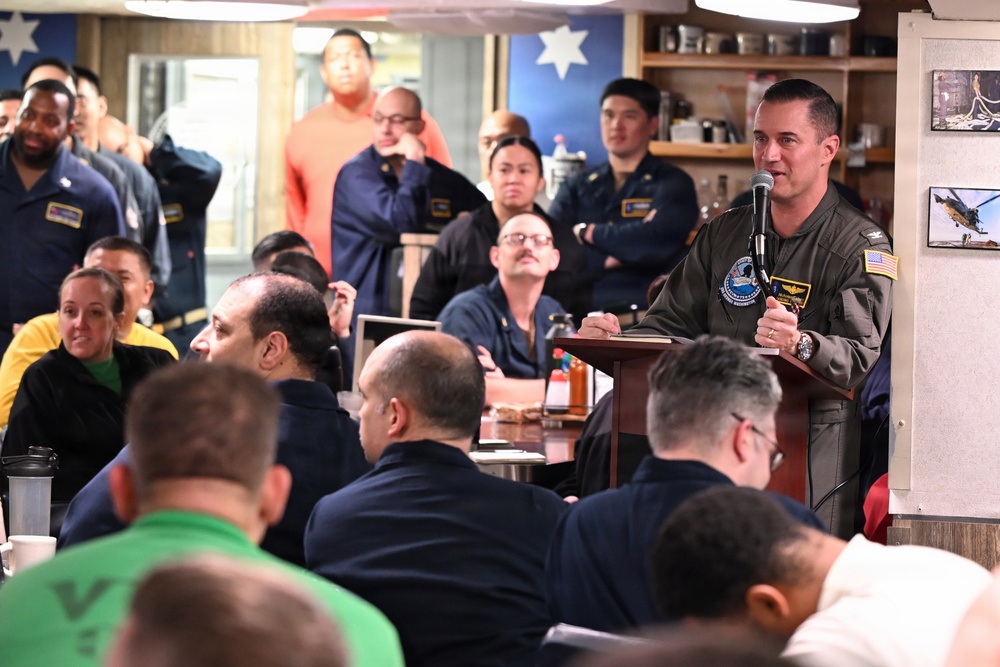 A military officer in an olive/sage green flight suit is addressing a group of personnel at a podium with a microphone in what appears to be a naval vessel's mess hall or briefing room. The audience consists of numerous uniformed military personnel seated and standing, listening attentively. The setting includes decorative elements such as a blue wall with a star, photographs on the wall, and condiment bottles on tables.