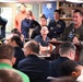 A military officer in an olive/sage green flight suit is addressing a group of personnel at a podium with a microphone in what appears to be a naval vessel's mess hall or briefing room. The audience consists of numerous uniformed military personnel seated and standing, listening attentively. The setting includes decorative elements such as a blue wall with a star, photographs on the wall, and condiment bottles on tables.