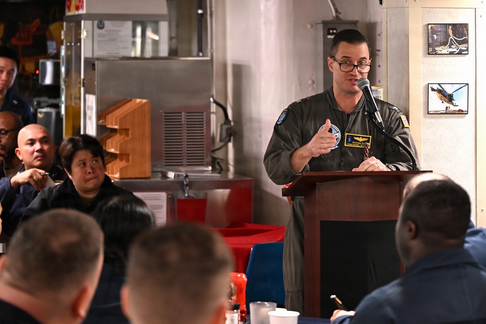 A military officer in a flight suit is speaking at a podium with a microphone, addressing a seated audience in what appears to be a military mess hall or briefing room aboard a naval vessel. The speaker wears glasses and a green flight suit with patches, including what appears to be a commanding officer designation. Several uniformed personnel are seated and listening attentively.