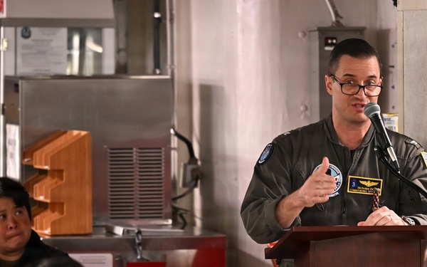 A military officer in a flight suit is speaking at a podium with a microphone, addressing a seated audience in what appears to be a military mess hall or briefing room aboard a naval vessel. The speaker wears glasses and a green flight suit with patches, including what appears to be a commanding officer designation. Several uniformed personnel are seated and listening attentively.