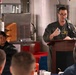 A military officer in a flight suit is speaking at a podium with a microphone, addressing a seated audience in what appears to be a military mess hall or briefing room aboard a naval vessel. The speaker wears glasses and a green flight suit with patches, including what appears to be a commanding officer designation. Several uniformed personnel are seated and listening attentively.