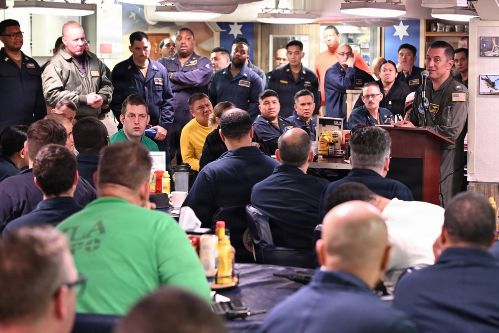 A military officer in a flight suit is addressing a large group of personnel in what appears to be a mess hall or common area aboard a naval vessel. The audience consists of numerous sailors and military personnel in various uniforms, seated and standing, attentively listening to the speaker at a podium. The setting suggests a formal briefing, all-hands meeting, or command address.