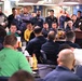 A military officer in a flight suit is addressing a large group of personnel in what appears to be a mess hall or common area aboard a naval vessel. The audience consists of numerous sailors and military personnel in various uniforms, seated and standing, attentively listening to the speaker at a podium. The setting suggests a formal briefing, all-hands meeting, or command address.