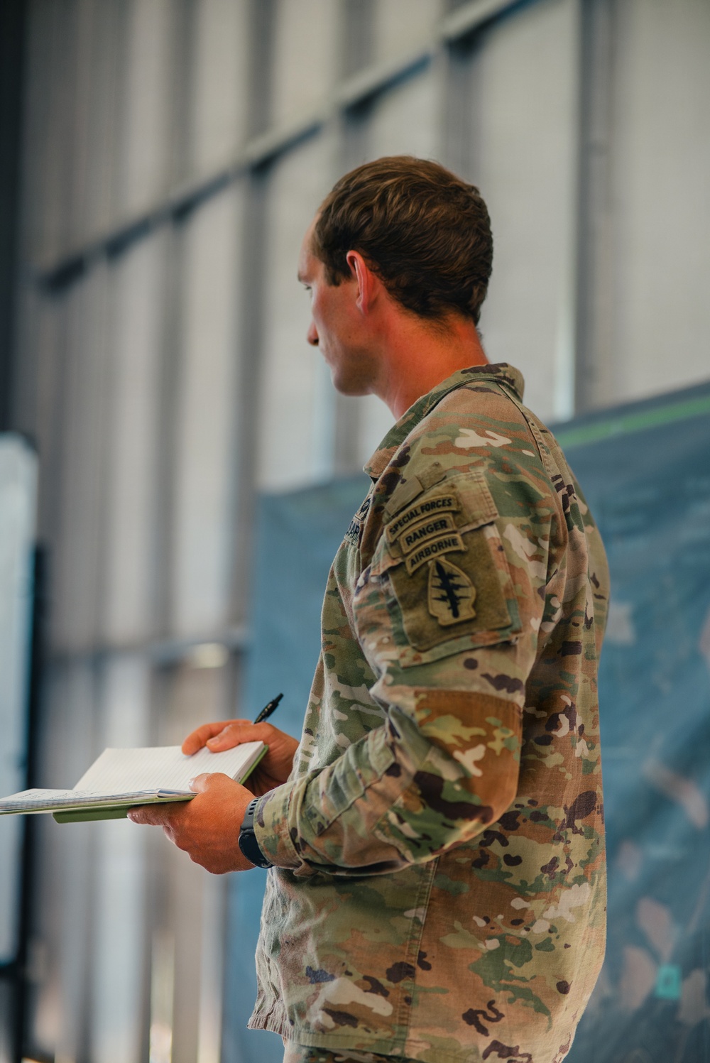 A U.S. Army soldier in Operational Camouflage Pattern (OCP) uniform is photographed from behind/side profile, holding a notebook and pen in what appears to be an indoor briefing or planning environment. The soldier displays multiple qualification tabs on the left shoulder sleeve, indicating an elite special operations background. A map or tactical display board is partially visible in the background.