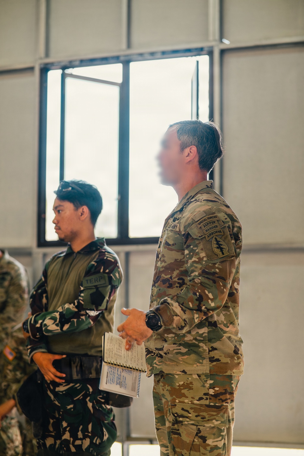 Two military personnel are standing indoors, appearing to attend a briefing or training session. The foreground figure wears US Army MultiCam (OCP) uniform with multiple qualification tabs, while the background figure wears a Philippine Armed Forces woodland camouflage uniform with a 'TERP' (interpreter) patch. This appears to be a joint US-Philippine military exercise or training event.