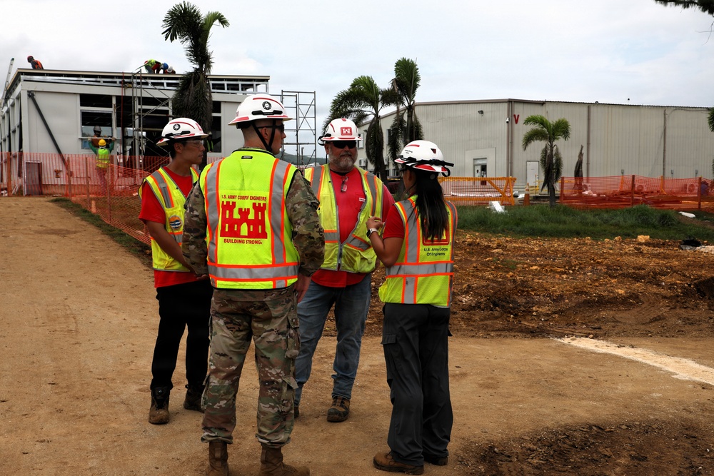 Four individuals are gathered at an active construction site, with at least three wearing U.S. Army Corps of Engineers high-visibility safety vests and white hard hats. One person is in military camouflage uniform also wearing a Corps of Engineers vest. They appear to be conducting a site inspection or project briefing in front of partially constructed buildings surrounded by orange safety fencing.