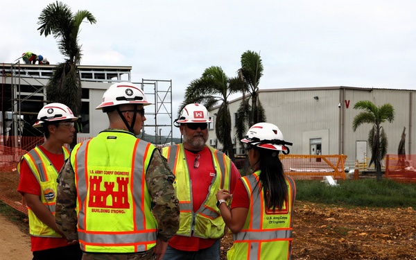 Four individuals are gathered at an active construction site, with at least three wearing U.S. Army Corps of Engineers high-visibility safety vests and white hard hats. One person is in military camouflage uniform also wearing a Corps of Engineers vest. They appear to be conducting a site inspection or project briefing in front of partially constructed buildings surrounded by orange safety fencing.