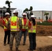 Four individuals are gathered at an active construction site, with at least three wearing U.S. Army Corps of Engineers high-visibility safety vests and white hard hats. One person is in military camouflage uniform also wearing a Corps of Engineers vest. They appear to be conducting a site inspection or project briefing in front of partially constructed buildings surrounded by orange safety fencing.
