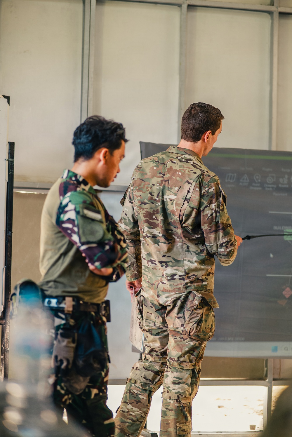 Two military personnel in different camouflage uniforms are standing before a briefing board or display screen. One soldier, wearing what appears to be a woodland/tiger-stripe pattern uniform, observes while the other, in a MultiCam/OCP pattern uniform, appears to be conducting or presenting a briefing using a pointer. The setting appears to be an indoor military facility or operations room.