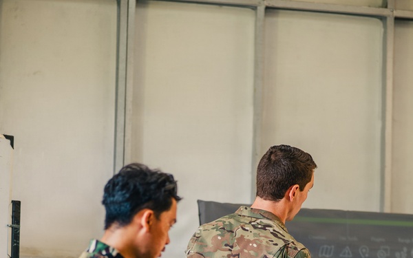Two military personnel in different camouflage uniforms are standing before a briefing board or display screen. One soldier, wearing what appears to be a woodland/tiger-stripe pattern uniform, observes while the other, in a MultiCam/OCP pattern uniform, appears to be conducting or presenting a briefing using a pointer. The setting appears to be an indoor military facility or operations room.