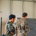 Two military personnel in different camouflage uniforms are standing before a briefing board or display screen. One soldier, wearing what appears to be a woodland/tiger-stripe pattern uniform, observes while the other, in a MultiCam/OCP pattern uniform, appears to be conducting or presenting a briefing using a pointer. The setting appears to be an indoor military facility or operations room.